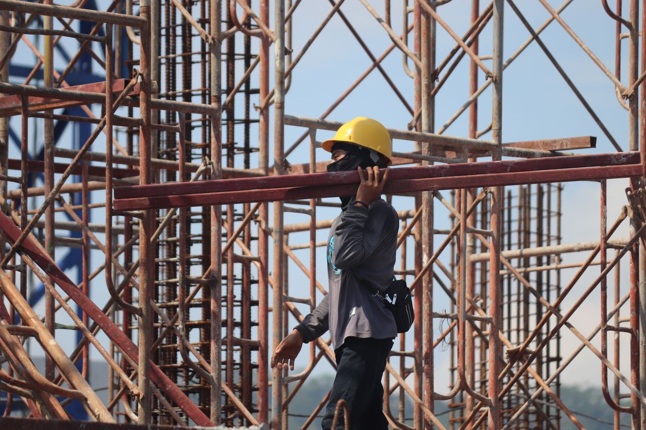 services-03 A construction worker with safety gear carrying a metal beam amidst scaffolding at a busy construction site.